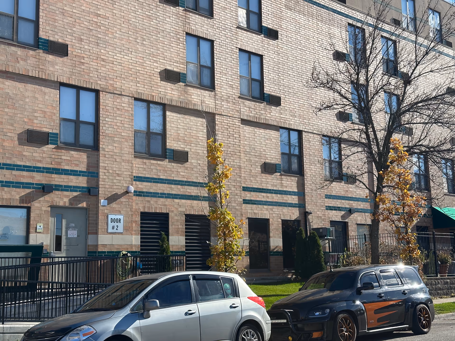 Exterior view of a brick building with multiple windows and a sign reading 'DOOR #2'. Two cars are parked on the street in front of the building, and there are small trees with autumn leaves near the sidewalk.