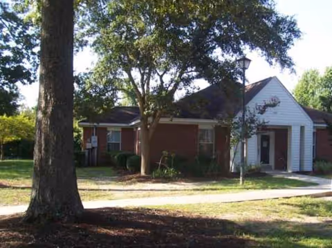 A single-story brick senior living facility with a white entrance, trees, lamp post, and sidewalk