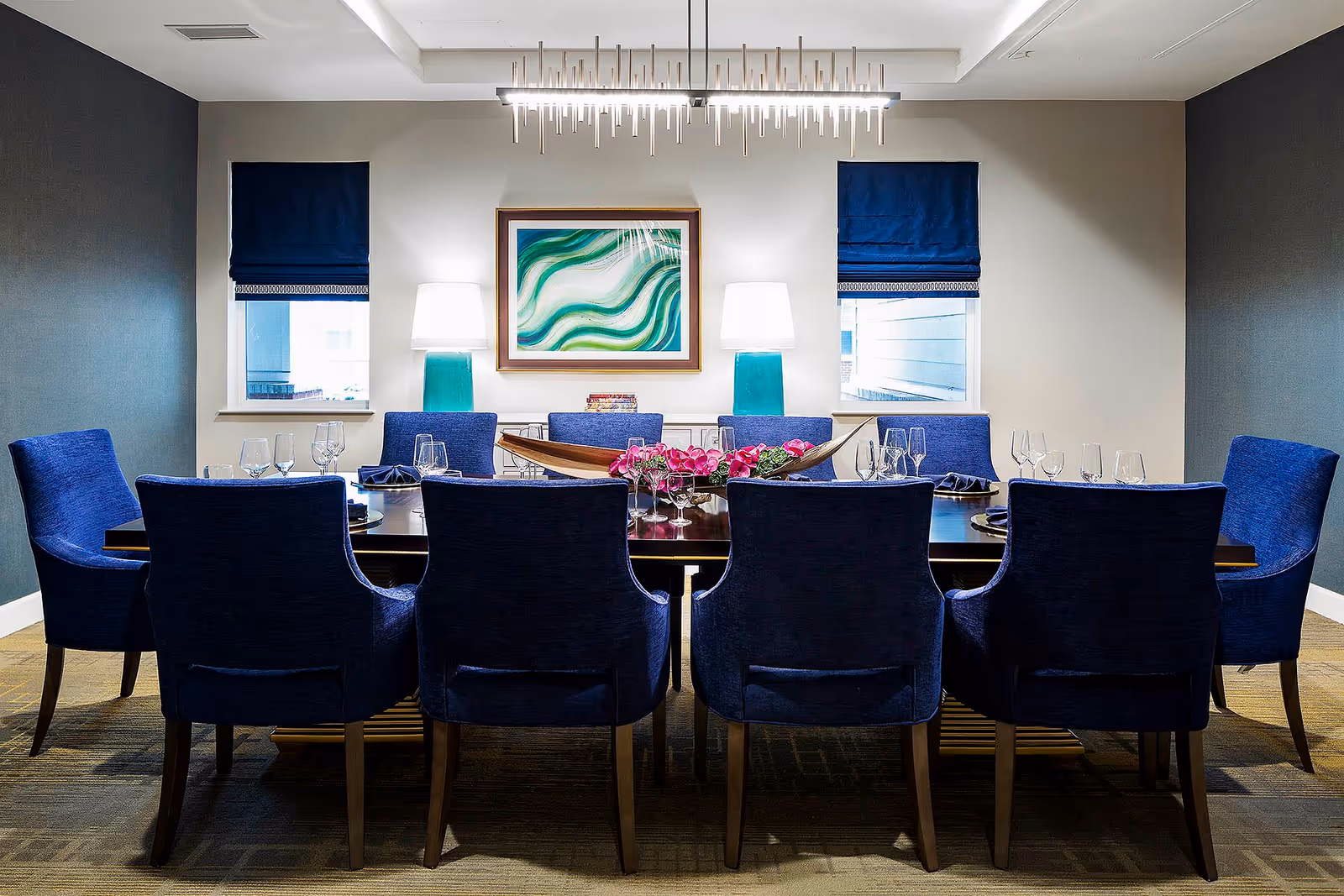 A modern dining room with a long dark wooden table surrounded by ten blue upholstered chairs. The table is set with wine glasses, plates, and napkins. Two windows with dark blue Roman shades flank a wall with a framed abstract green and white painting. Two turquoise table lamps with white shades are placed on a sideboard beneath the painting. A contemporary chandelier hangs above the table.