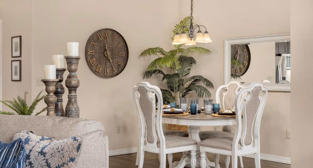 A bright dining area with a round table and four upholstered chairs, decorative wall clock, potted plants and a hanging chandelier next to a living room sofa.