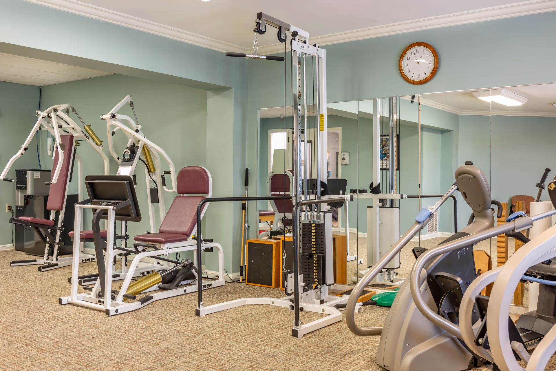 Exercise room with weight machines, cardio equipment, mirrors and a wall clock.