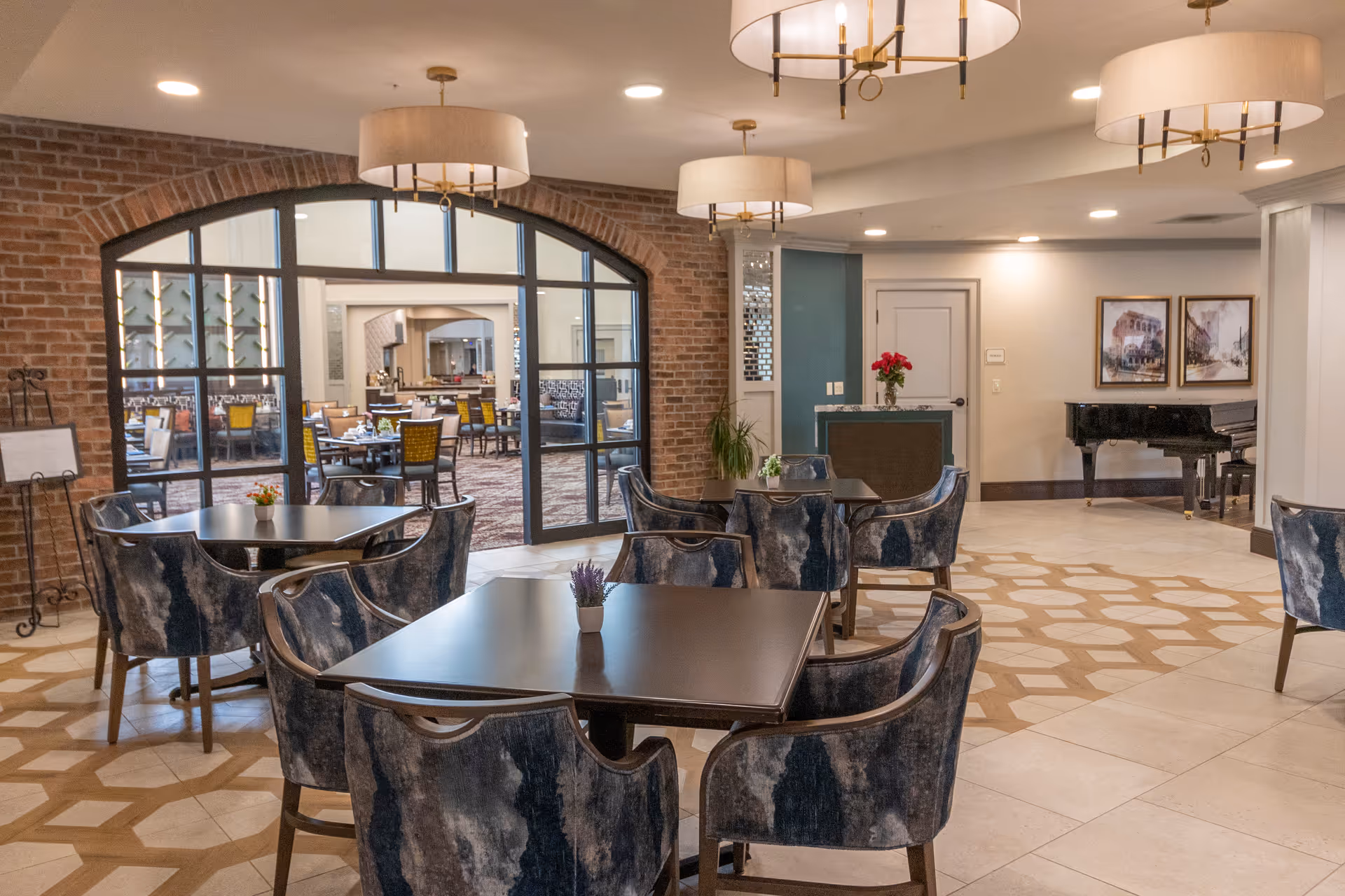 A spacious and well-lit common area with several dark wood tables surrounded by blue and gray upholstered chairs. The floor features a geometric tile pattern. In the background, there is a brick archway with glass panels leading to a dining room with more tables and chairs. The room also includes a black grand piano, framed artwork on the walls, and modern ceiling light fixtures.