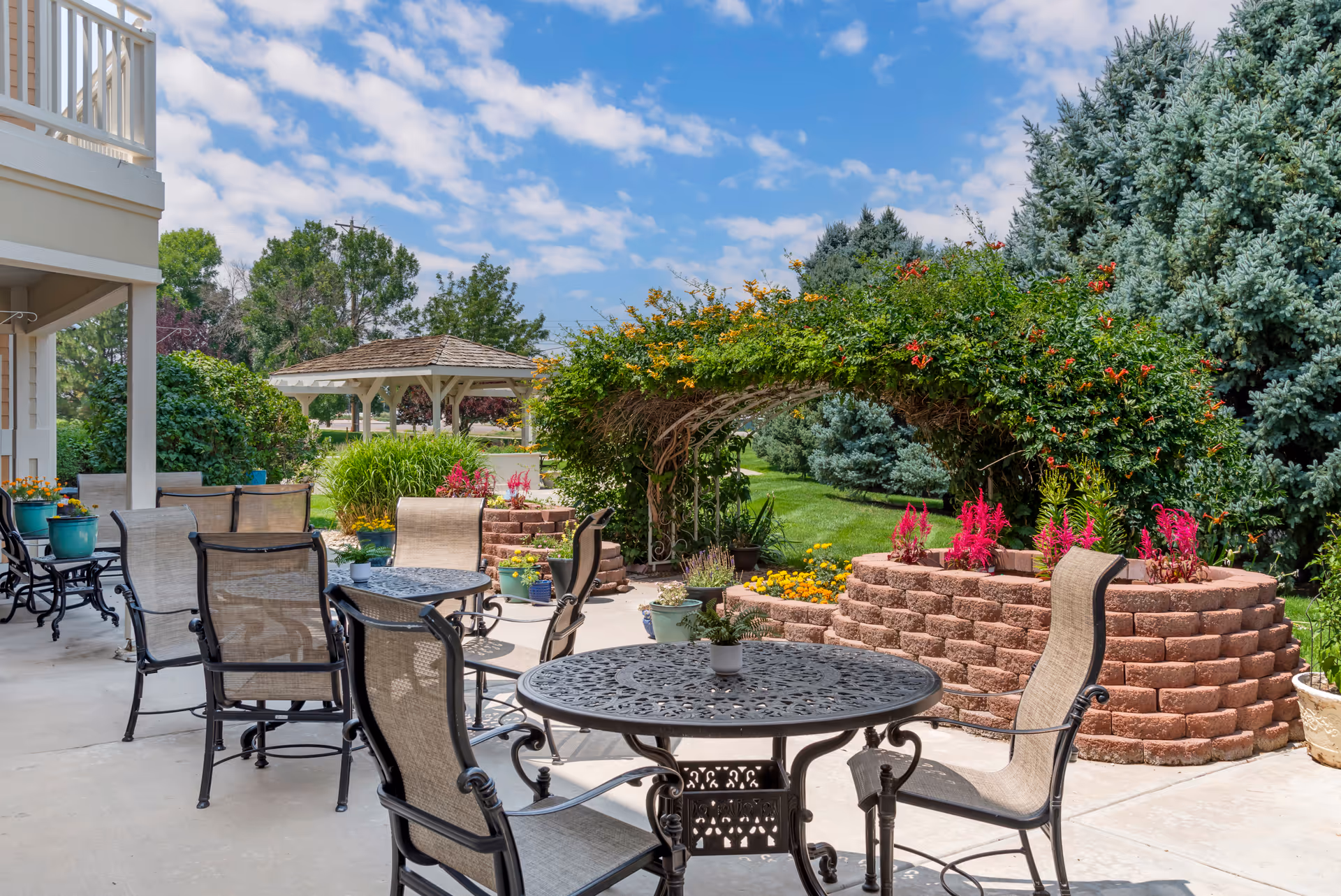 Outdoor patio area with metal tables and chairs, surrounded by lush greenery, colorful flowers, a vine-covered archway, and a gazebo in the background under a partly cloudy sky.