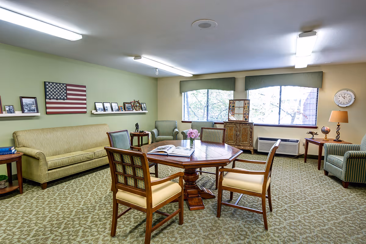 A cozy living room with a patterned carpet, a wooden octagonal table surrounded by four cushioned chairs, a beige sofa against a green wall decorated with framed photos and an American flag, two striped armchairs near a window with green valances, a wooden cabinet, and side tables with lamps and decorative items.