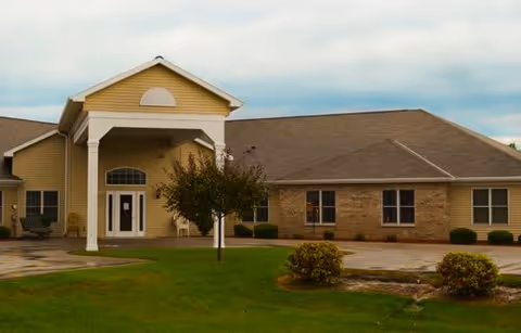 Exterior view of Harbor View Assisted Living facility showing a single-story building with a covered entrance supported by white columns, beige siding, brick accents, and a well-maintained lawn with small bushes and a tree in front.