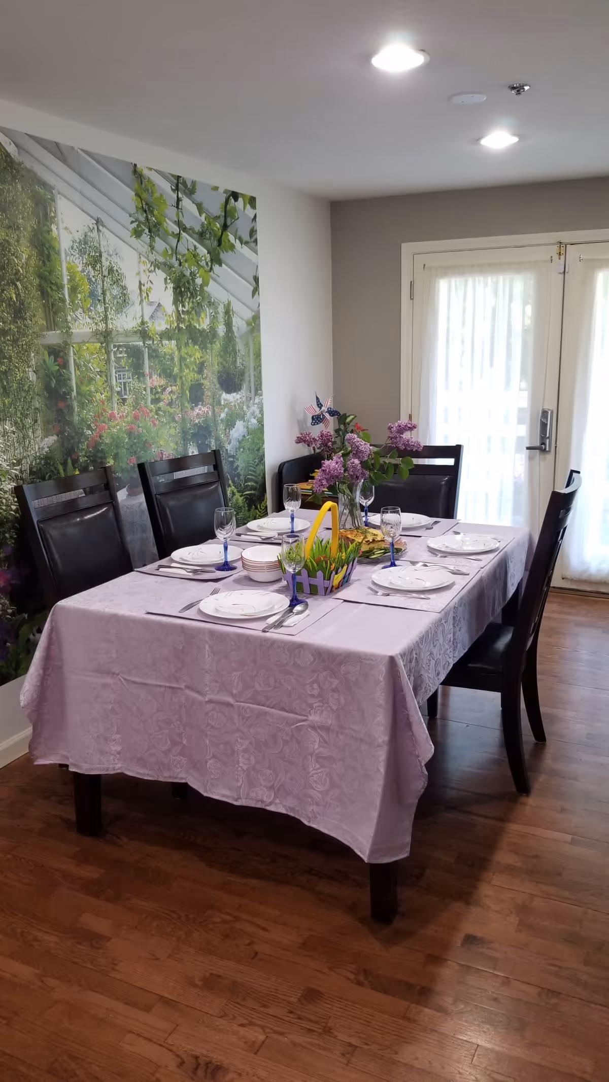 A dining room with a rectangular table covered with a lavender tablecloth, set with white plates, bowls, silverware, and blue-stemmed glasses. There is a centerpiece with purple flowers and a small decorative basket with yellow and green accents. The room has wooden flooring, black dining chairs, a large wall mural of a garden scene, and glass double doors with sheer white curtains letting in natural light.
