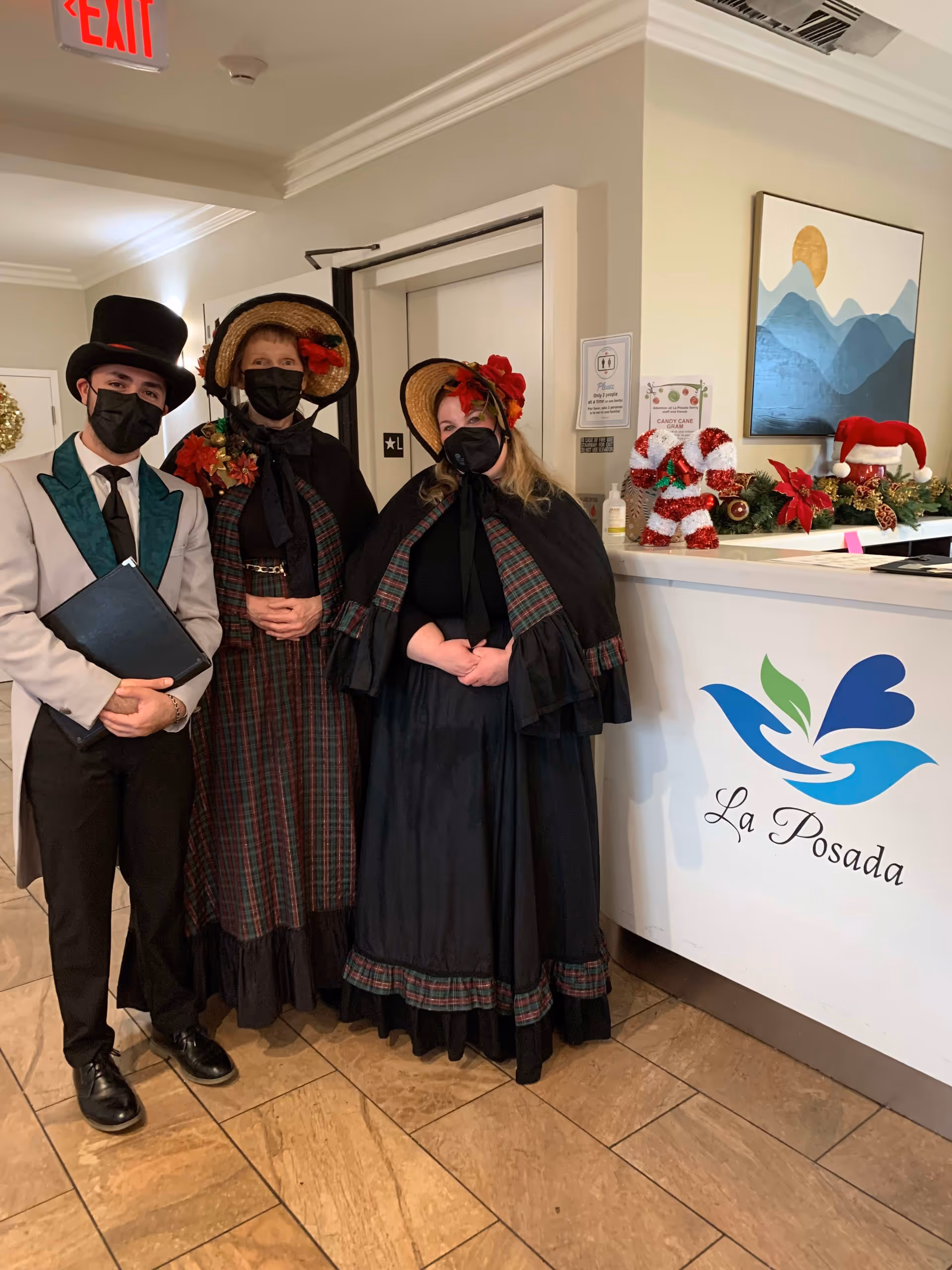 Three people dressed in Victorian-style clothing and black face masks standing inside a facility near a reception desk decorated with Christmas ornaments and a sign that reads La Posada. The man on the left wears a white jacket with green lapels and a black top hat, holding a black folder. The two women wear matching plaid dresses with black shawls and straw bonnets adorned with red flowers.