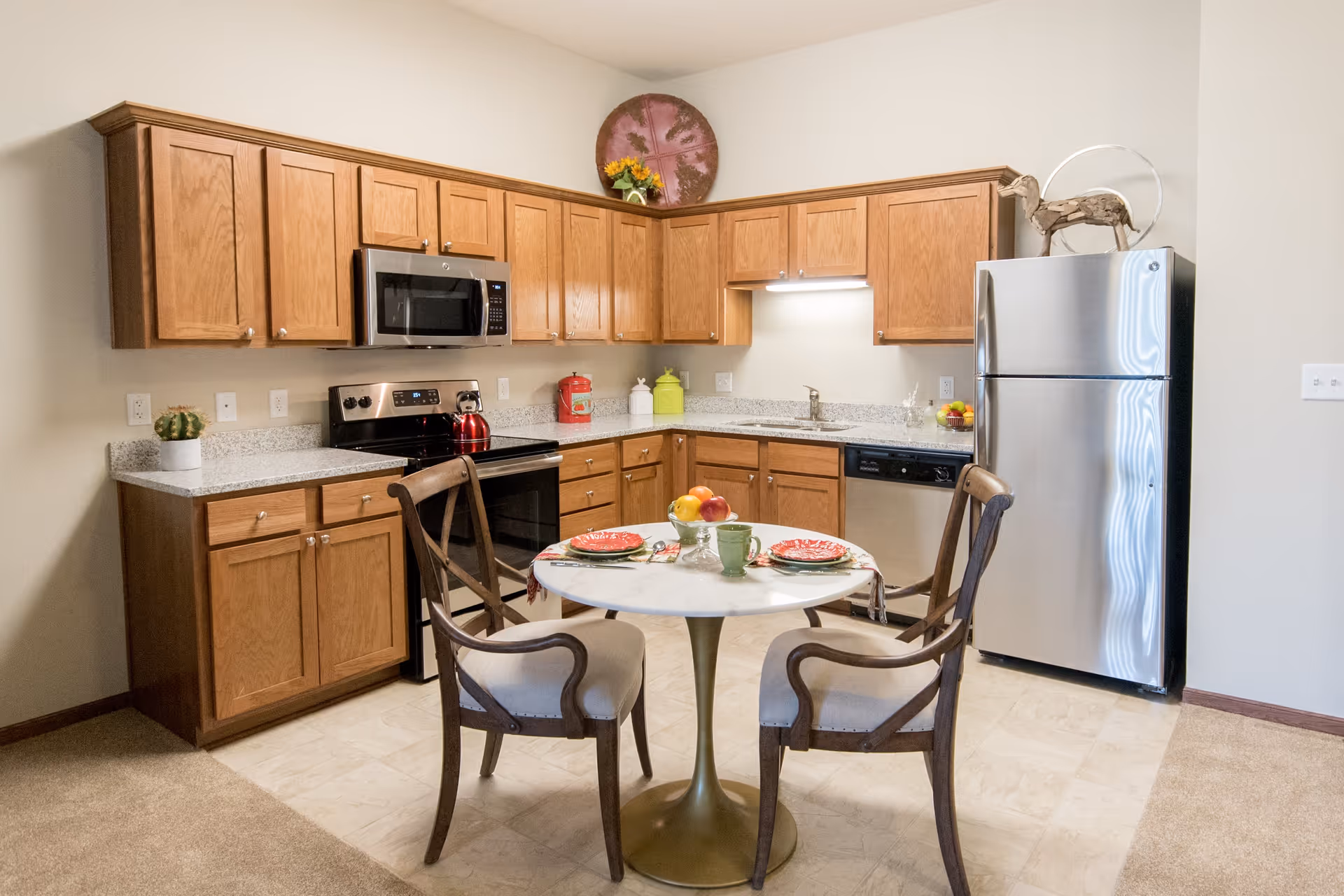 A kitchen area with wooden cabinets, a stainless steel refrigerator, microwave, stove, and dishwasher. In the center, there is a small round dining table set for two with plates, cups, and a bowl of fruit. The floor is a combination of tile and carpet, and decorative items are placed on top of the cabinets and refrigerator.