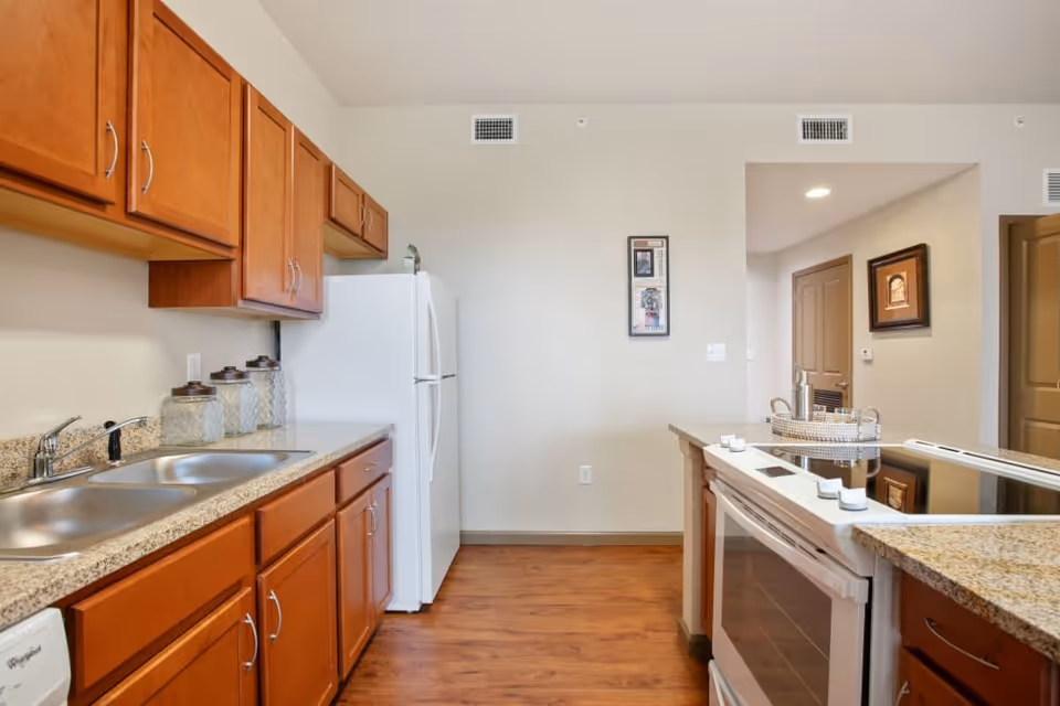 A clean and modern kitchen with wooden cabinets, a double stainless steel sink, a white refrigerator, and a stove with a smooth black cooktop. The kitchen has granite countertops and wood flooring. There are decorative jars on the counter and framed artwork on the walls.