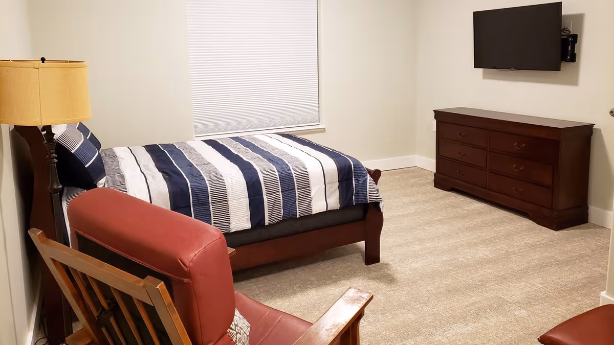 Neatly furnished bedroom with a bed in striped bedding, a dresser with a wall-mounted TV, and a red armchair.