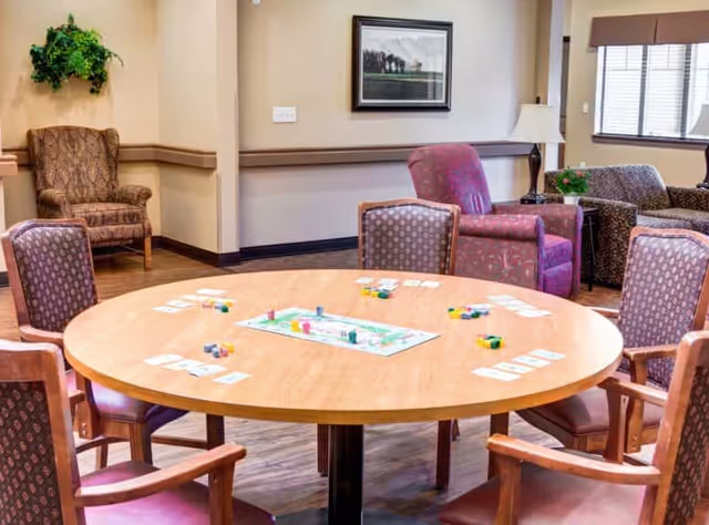 Round activity table set up for a board game surrounded by upholstered chairs in a furnished memory care common room.