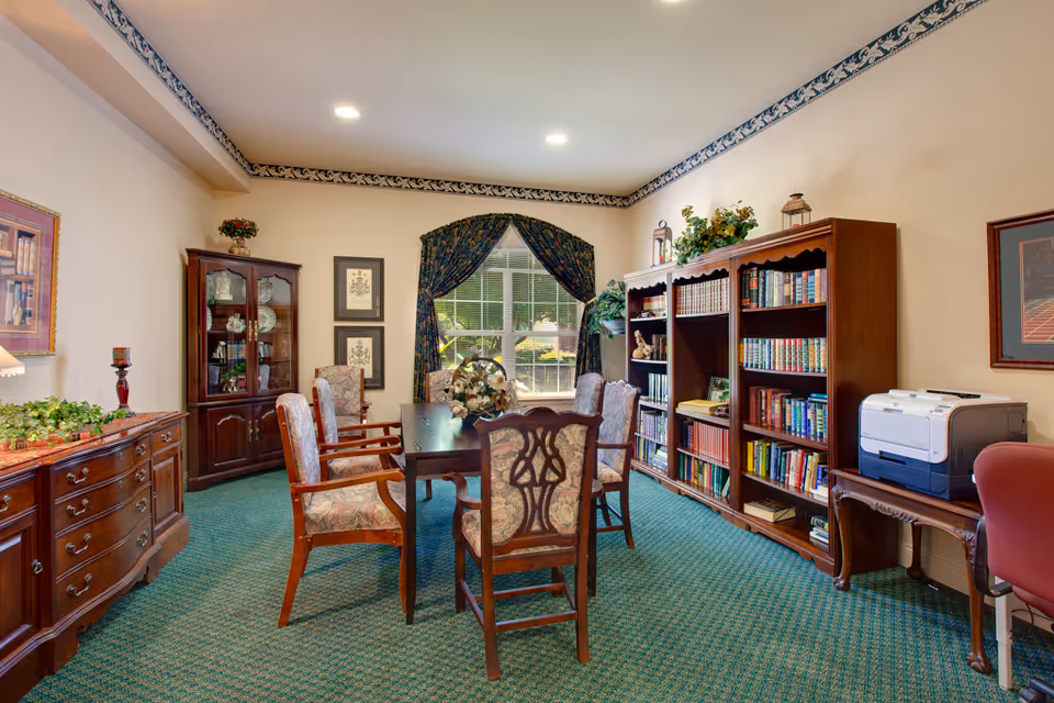 A cozy room with a wooden table surrounded by six upholstered chairs. The room features a green carpet, a large window with dark floral curtains, wooden bookshelves filled with books, a wooden cabinet with glass doors displaying decorative items, and a wooden sideboard with plants and a candle. There is also a printer on a small wooden table and framed artwork on the walls.