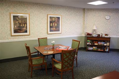 A cozy common area with a square wooden table surrounded by four green cushioned chairs. On the table are some magazines and a small vase with white flowers. The walls are decorated with two framed paintings and a clock. In the corner, there is a wooden bookshelf with various board games and a lamp on top.