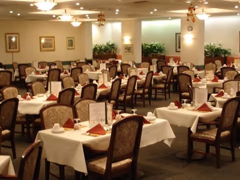 A spacious dining room with multiple tables covered in white tablecloths, each set with cups, plates, and folded red napkins. The room has upholstered chairs, framed artwork on the walls, ceiling lights, and plants placed around the space.