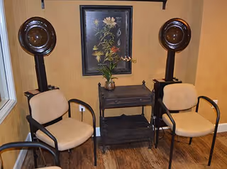 Two beige salon chairs with black armrests positioned in front of two vintage hair dryers mounted on stands. Between the chairs is a small dark wooden table with a vase of flowers on top. A framed floral painting hangs on the wall behind the setup. The room has wooden flooring and light brown walls.