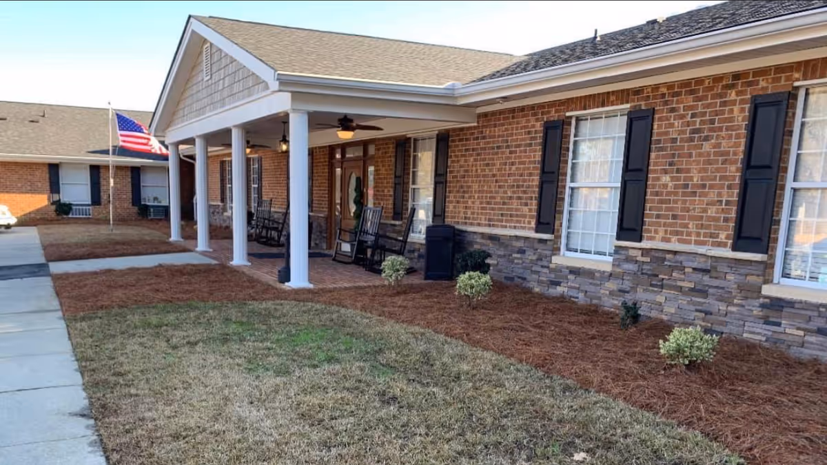 Brick senior living facility entrance with a covered porch, white columns, rocking chairs, and an American flag on the lawn.