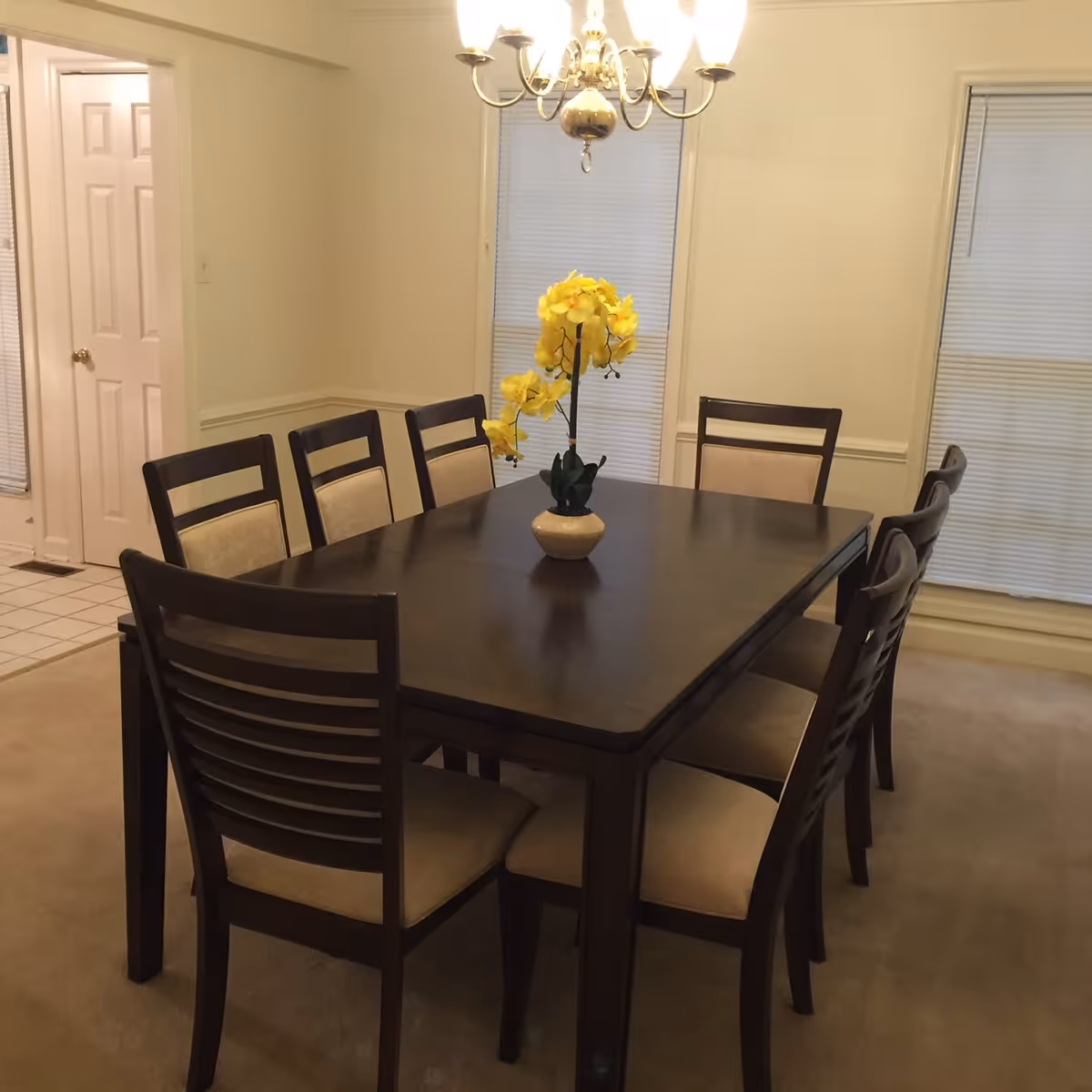 A dining room with a dark wooden rectangular table surrounded by eight matching chairs with beige cushions. A vase with yellow flowers is placed in the center of the table. The room has beige walls, two windows with closed blinds, a chandelier hanging above the table, and a door leading to another room with tiled flooring.