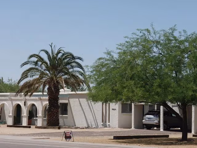 Exterior view of a single-story white building with arched doorways, a large palm tree, and another leafy tree. A car is parked under a covered area on the right side, and a small sign is placed on the ground near the front.