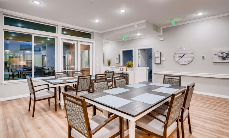 A bright and clean dining room with two rectangular tables, each surrounded by six chairs with patterned backs and white cushions. The tables have light blue placemats. Large windows and glass doors reveal an outdoor seating area. The walls are light gray with white trim, decorated with a large clock and a floral painting. The floor is light wood, and there are green exit signs above two doorways in the background.