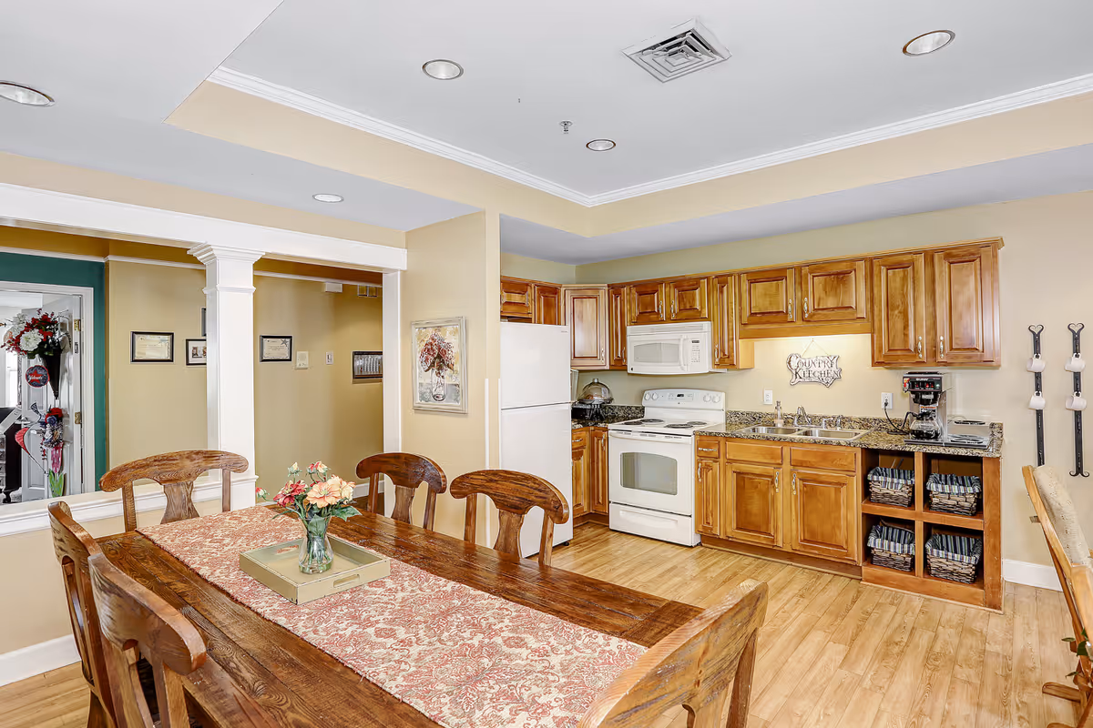 A cozy kitchen and dining area featuring wooden cabinets, a white refrigerator, stove, and microwave. The dining table is wooden with a floral table runner and a vase of flowers in the center. The walls are painted beige, and the floor is wood. There is a decorative sign on the kitchen wall that reads 'Country Kitchen'.