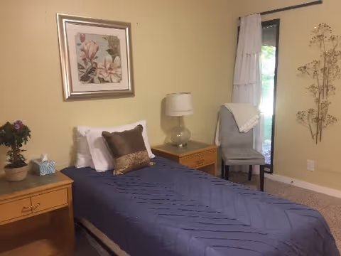 A small bedroom with a single bed covered in a blue quilt and two pillows. There are two wooden nightstands on either side of the bed, one with a potted plant and tissue box, the other with a glass lamp. A framed floral picture hangs on the wall above the bed. A gray upholstered chair with a white throw is placed near a window with white curtains. The walls are painted light yellow and the floor is carpeted.