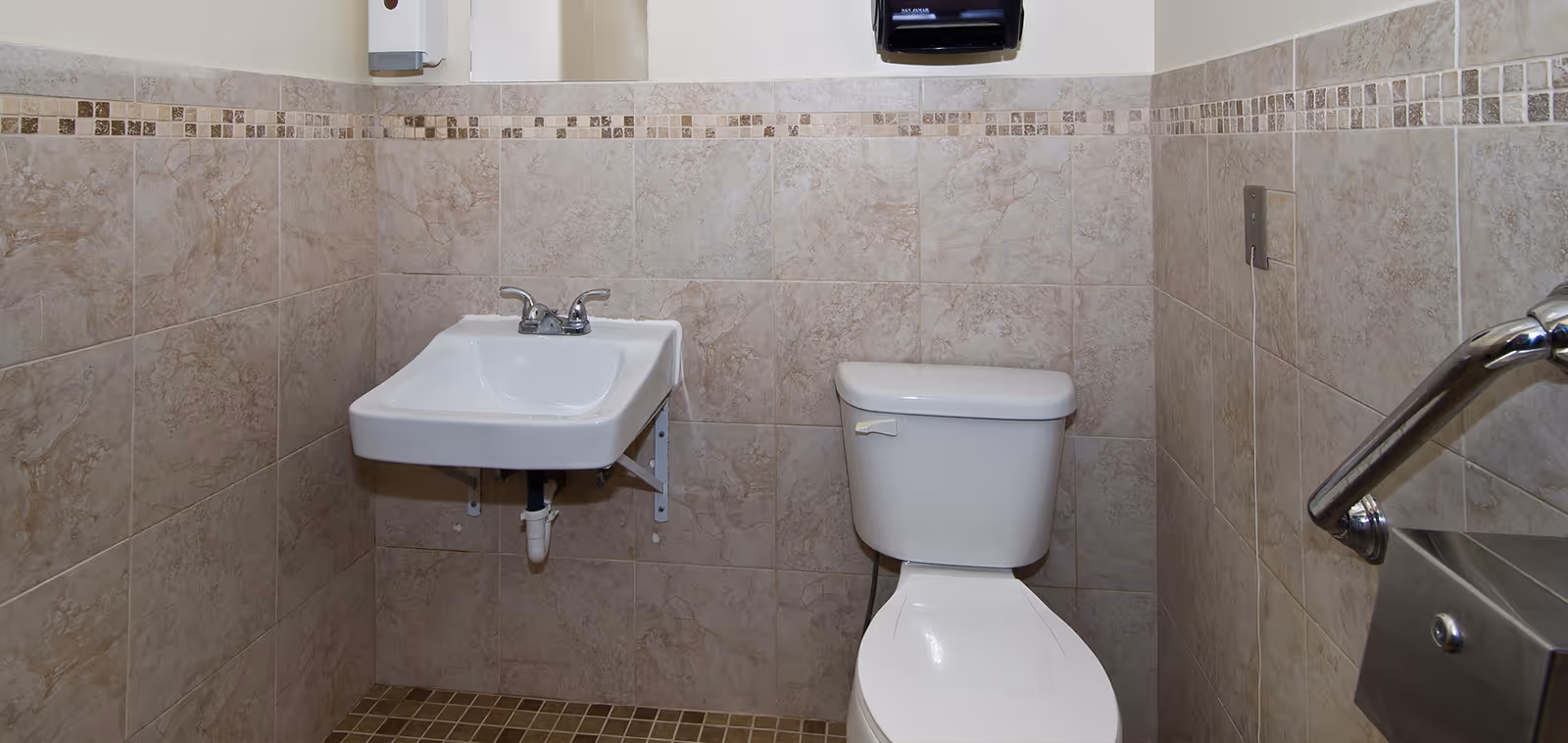 A small bathroom with beige tiled walls and floor, featuring a white wall-mounted sink with a faucet, a white toilet, a metal grab bar on the right wall, a soap dispenser above the sink, and a paper towel dispenser on the wall above the toilet.
