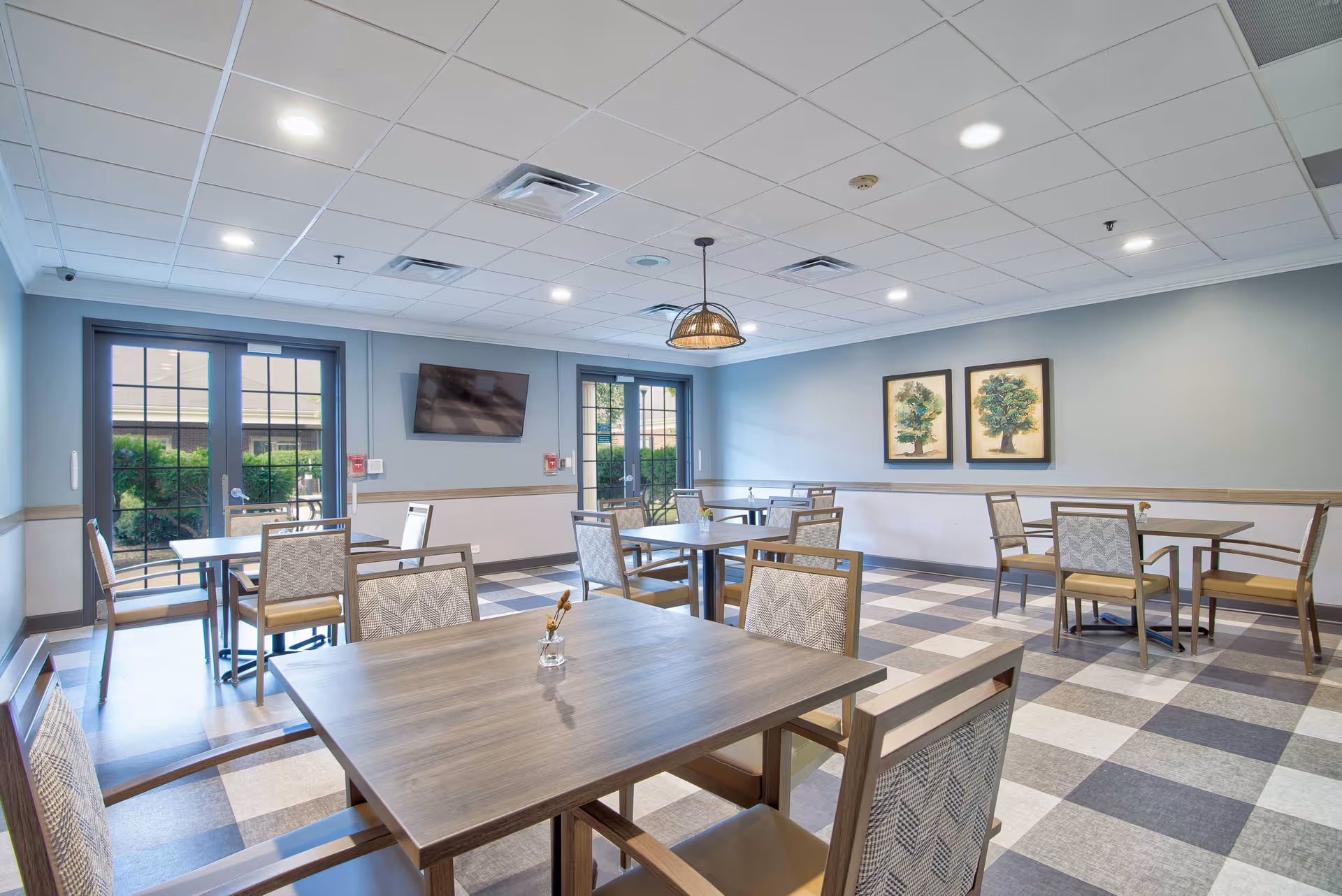 A bright dining room with multiple square wooden tables and cushioned chairs arranged neatly. The room has large windows and glass doors letting in natural light, a wall-mounted TV, two framed tree artworks on the wall, and a checkered tile floor.