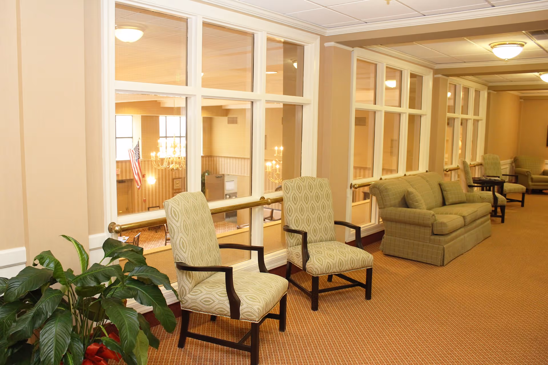 A well-lit hallway in a senior living facility with large windows overlooking a lower level. The hallway features patterned armchairs, a green sofa, side tables, and a potted plant. The walls are beige with white trim, and the carpet is a warm brown color.