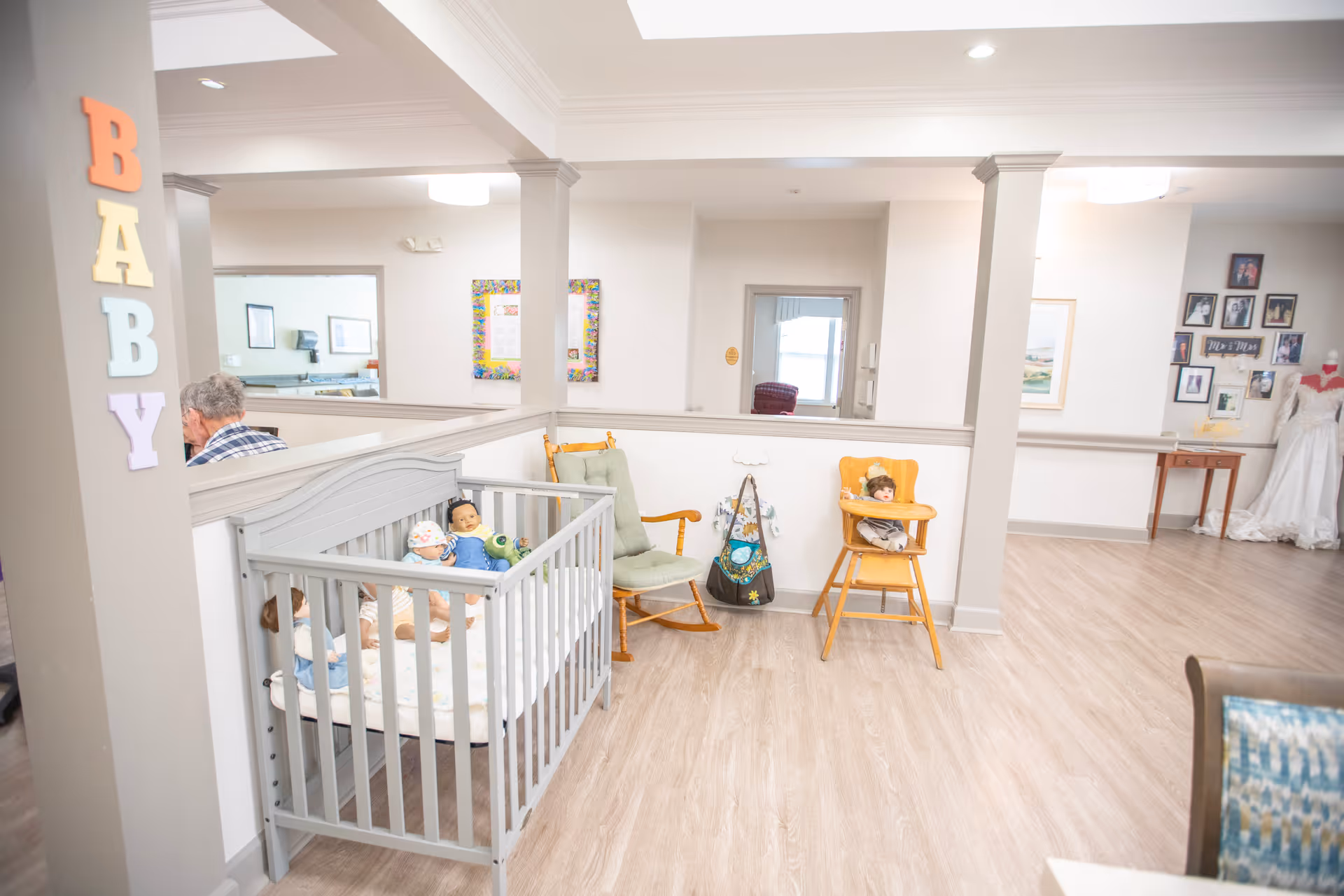 Interior of a senior living facility area with a gray baby crib containing dolls, a green rocking chair, a wooden high chair with a doll, and the word BABY spelled out in colorful letters on a pillar. There are framed pictures on the walls and a wedding dress displayed on a mannequin in the background.