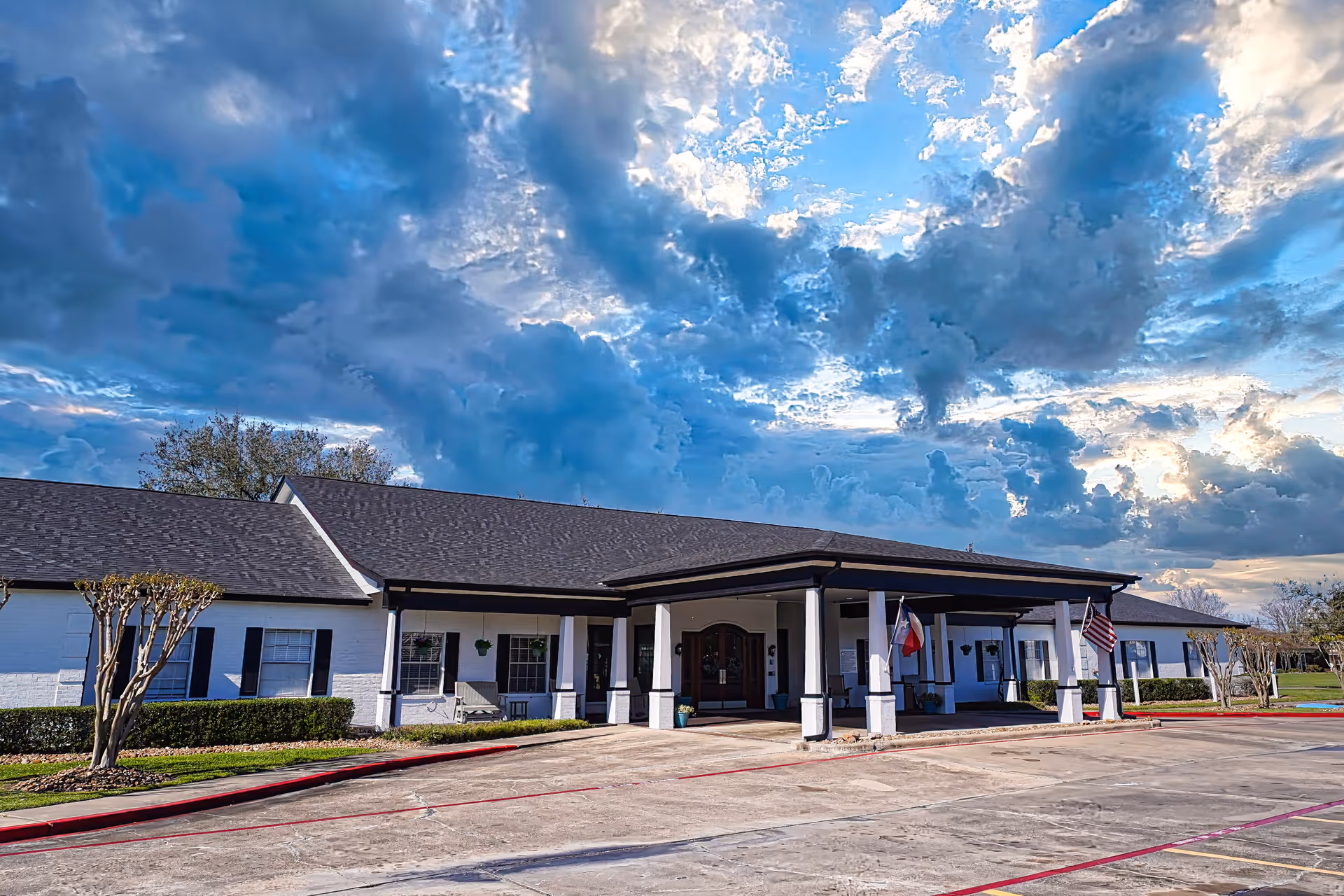 Exterior view of a single-story building with a covered entrance supported by white columns. The building has white walls with black shutters and a dark roof. There are two flags displayed near the entrance, and the sky above is filled with dramatic clouds during sunset or sunrise.
