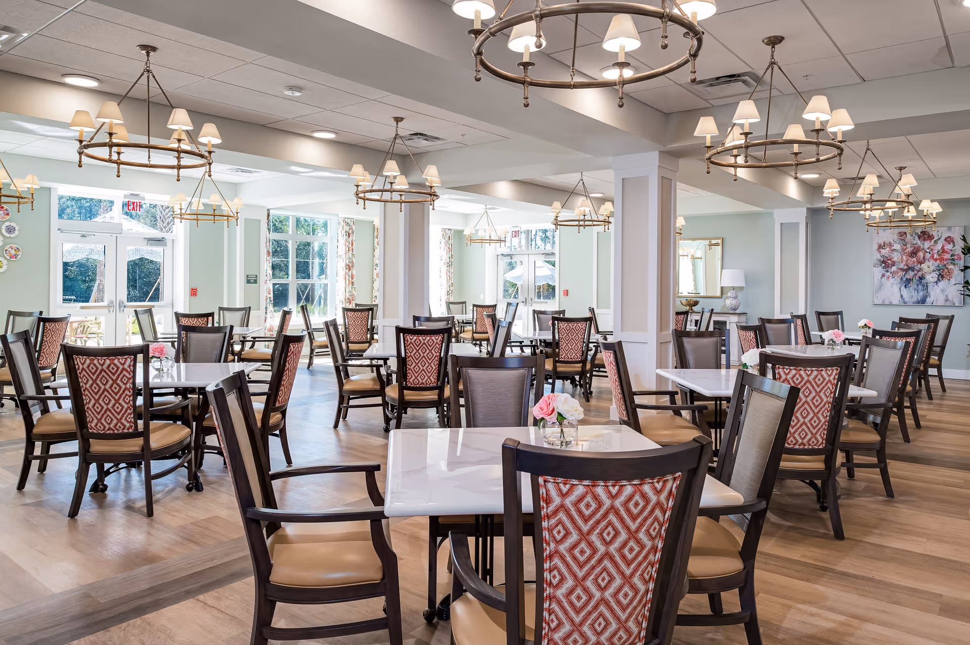 A bright and spacious dining room with multiple square tables and wooden chairs featuring patterned red and beige upholstery. The room has large windows with floral curtains allowing natural light to fill the space. Elegant circular chandeliers with multiple lampshades hang from the ceiling. There are small flower arrangements on each table and a large floral painting on the far wall.