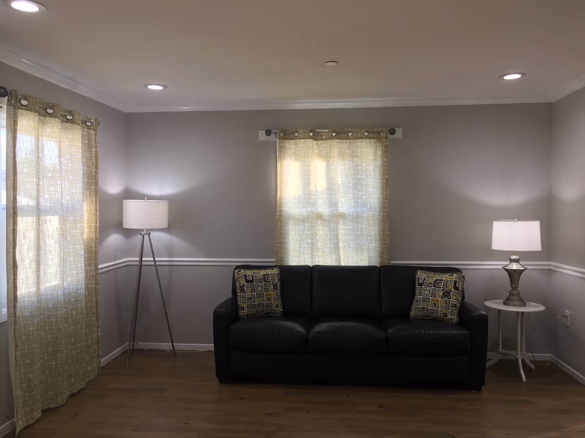 A living room with a black leather sofa adorned with two patterned cushions. There are two windows with light-colored curtains allowing natural light to filter through. On the left side of the sofa is a tall floor lamp with a white shade, and on the right side is a small round white table with a silver table lamp. The walls are painted light gray with white trim, and the floor is wooden.