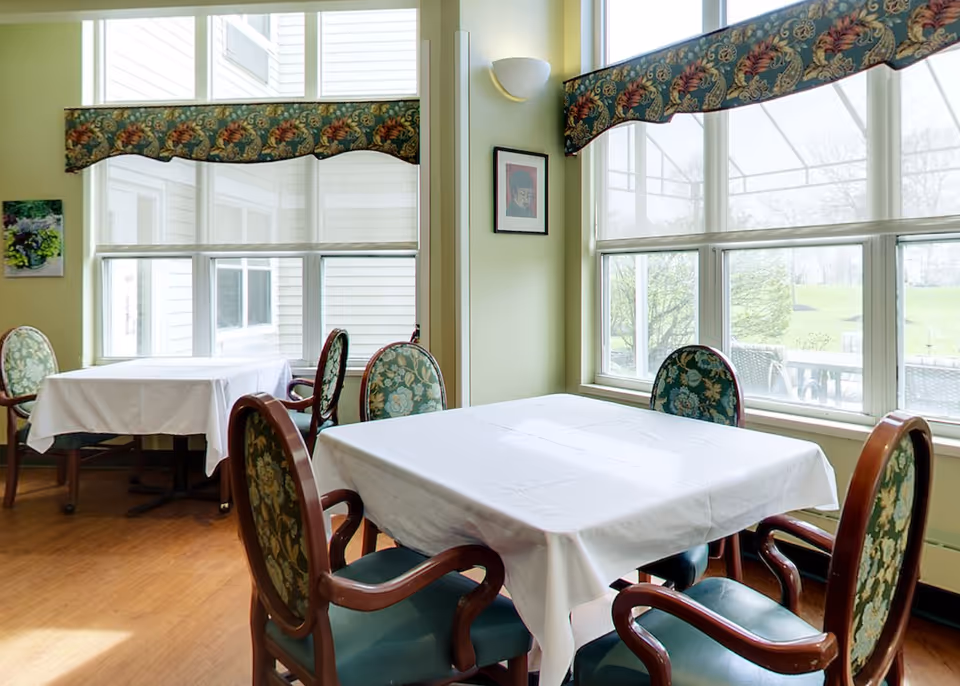 A bright dining area with two tables covered in white tablecloths, each surrounded by four wooden chairs with floral upholstered backs and seats. Large windows with floral valances let in natural light and offer a view of greenery outside. The walls are painted light green and decorated with framed artwork and a wall sconce.