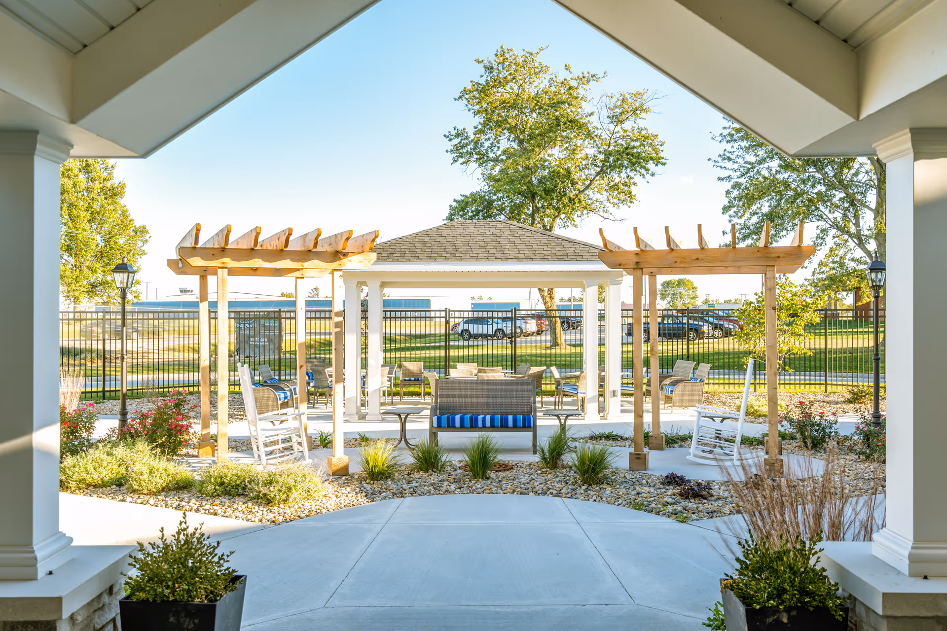 View from a covered patio looking out onto an outdoor seating area with wooden pergolas, rocking chairs, cushioned benches, and a gazebo surrounded by plants and trees under a clear blue sky.