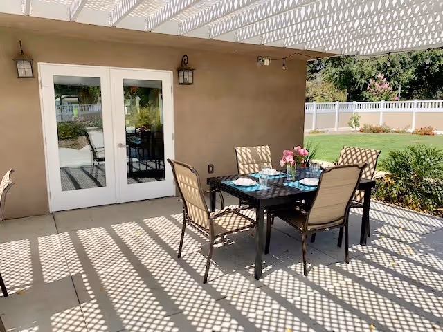 Outdoor patio area with a black metal table set for four with plates and glasses, surrounded by four beige cushioned chairs. The patio is shaded by a white lattice pergola casting patterned shadows on the concrete floor. There are double glass doors leading inside, and a white fence with greenery and lawn in the background.
