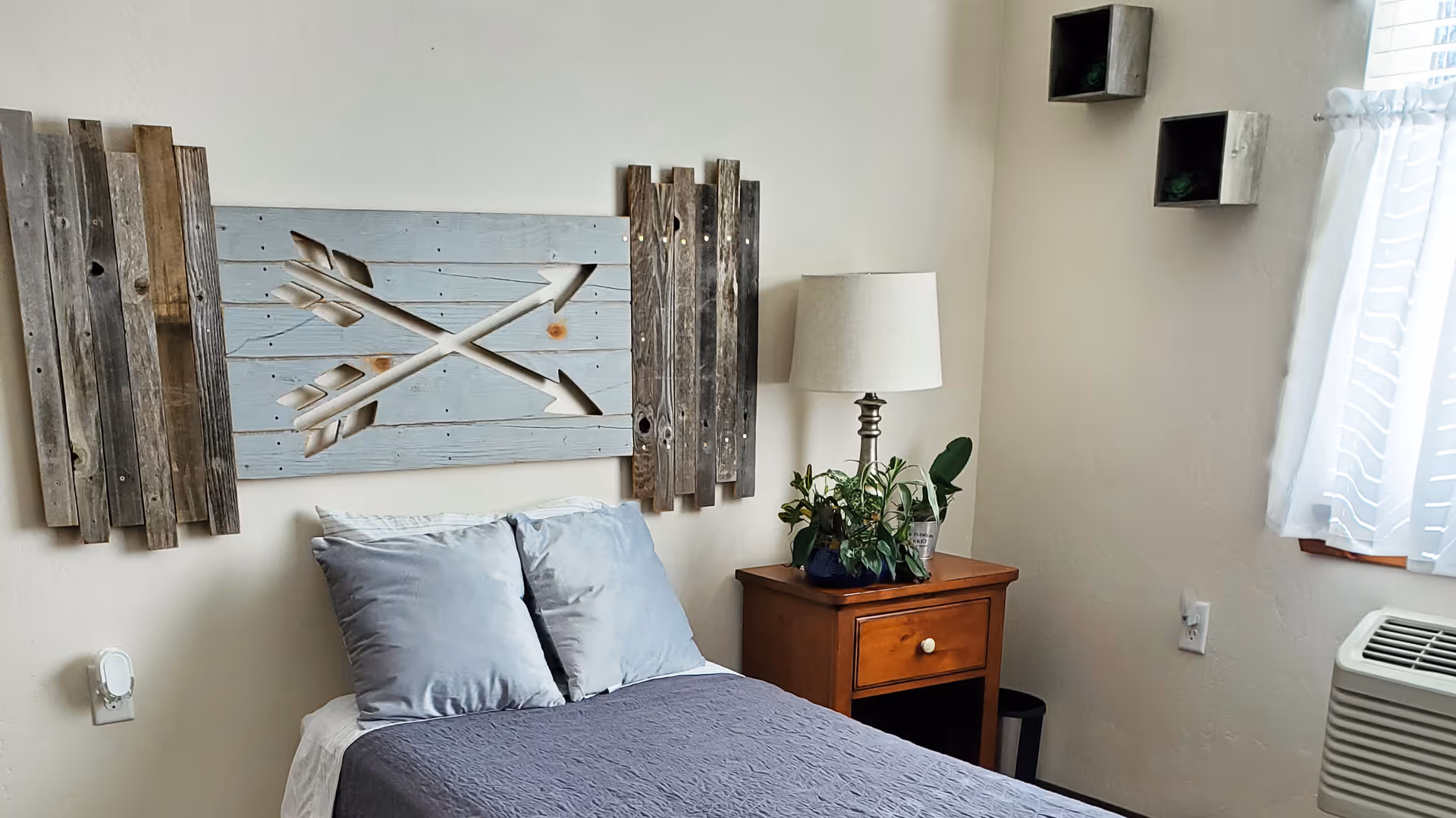 A cozy bedroom with a bed featuring gray pillows and a dark gray blanket. Above the bed is a decorative wooden wall art with two crossed arrows. Next to the bed is a wooden nightstand with a lamp and a potted plant. A window with sheer white curtains allows natural light into the room.