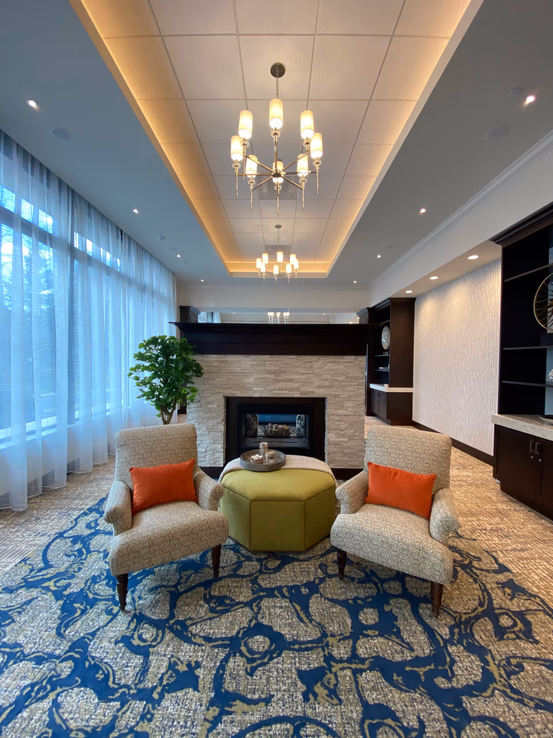 A cozy sitting area in a senior living facility featuring two upholstered armchairs with orange cushions, a green hexagonal ottoman with a tray on top, a modern fireplace with a stone surround, large windows with sheer curtains, a patterned blue and beige carpet, and elegant ceiling chandeliers.