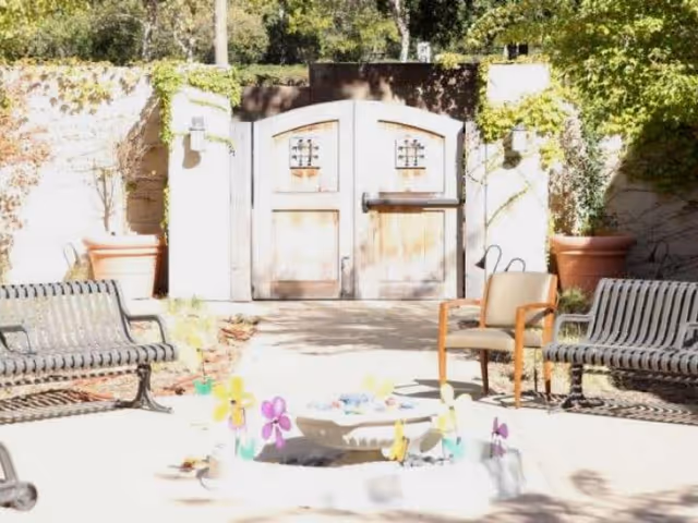 Sunlit outdoor courtyard with benches, a chair, potted plants and a wooden double gate, centered around a small decorative fountain with colorful pinwheel flowers.