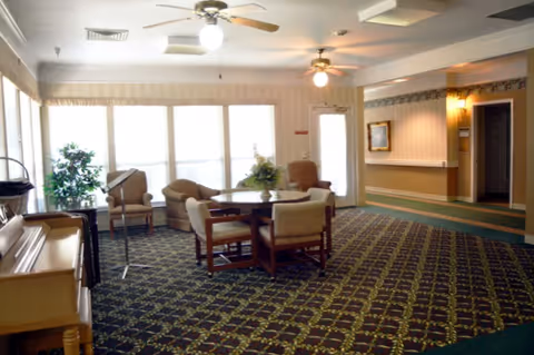 A spacious common area in an assisted living facility with patterned carpet, a round table surrounded by four chairs, two armchairs near large windows with blinds, a piano on the left, ceiling fans with lights, and a hallway leading to other rooms.