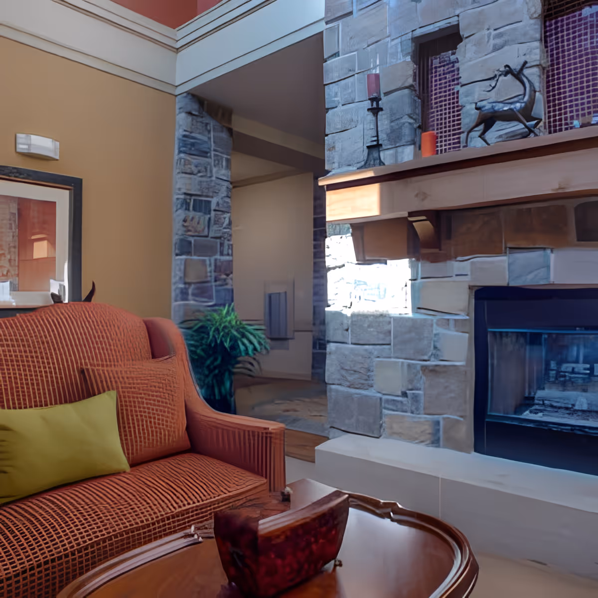 Cozy living room area featuring a red textured sofa with a green pillow, a wooden coffee table, and a stone fireplace adorned with decorative items including a metal deer sculpture and candles. The room has warm beige walls and a framed picture on the wall.