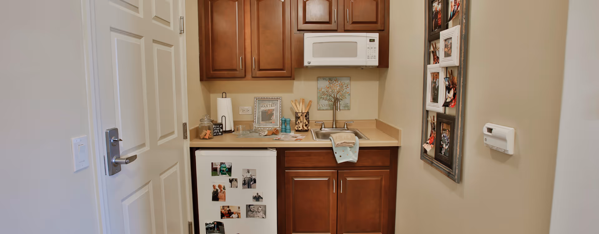 Small kitchenette with wooden cabinets, a white microwave mounted above the counter, a small refrigerator with photos attached, a sink with a towel draped over the edge, and various kitchen items on the countertop. A door is partially open on the left, and a photo frame with multiple pictures hangs on the right wall.