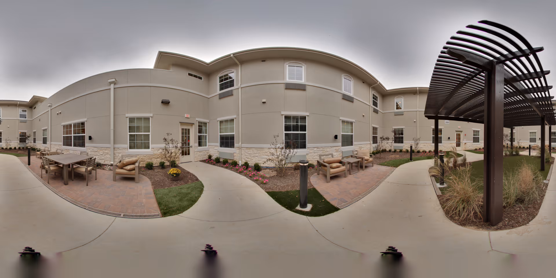 Outdoor courtyard area of a senior living facility with paved walkways, seating areas including chairs and tables, landscaped flower beds, and a wooden pergola structure. The building exterior is beige with multiple windows and doors.