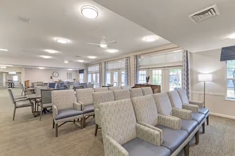 A spacious, well-lit common area in an assisted living facility featuring multiple rows of cushioned chairs with armrests arranged in the foreground. In the background, there are several tables and chairs near large windows with curtains, allowing natural light to fill the room. The ceiling has multiple round light fixtures and a ceiling fan, and a floor lamp is visible near a window on the right side.