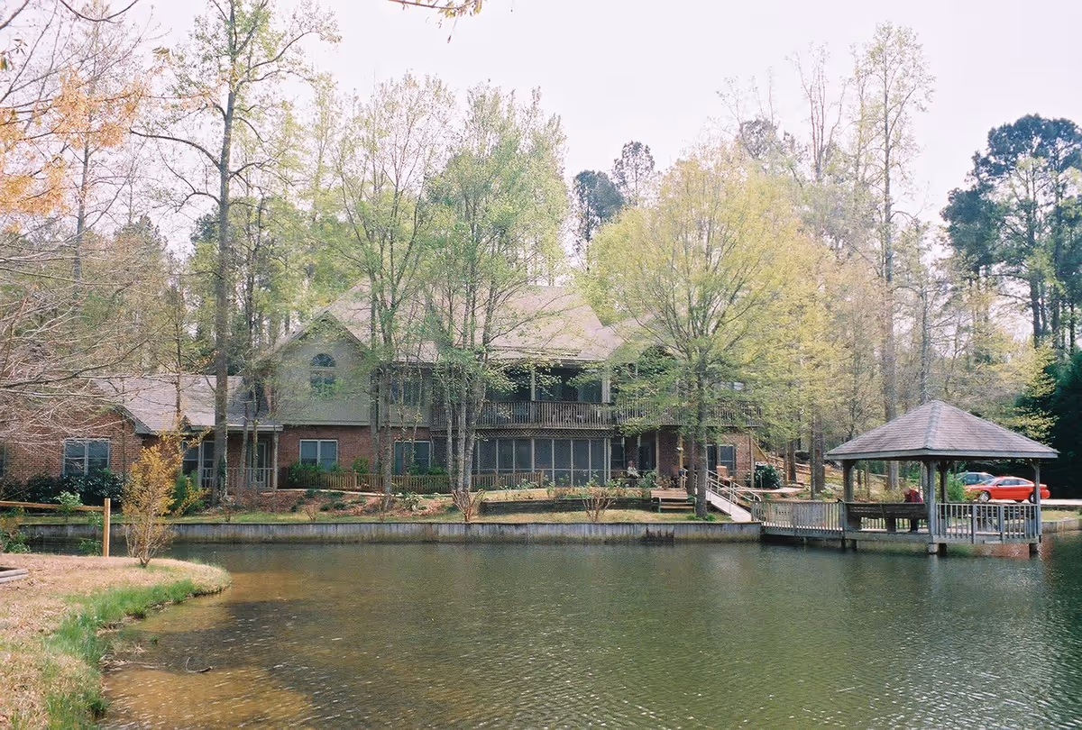 A large two-story brick building partially obscured by trees, situated beside a calm pond with a wooden gazebo on a dock extending over the water. A red car is parked near the gazebo, and the scene is surrounded by greenery and trees.