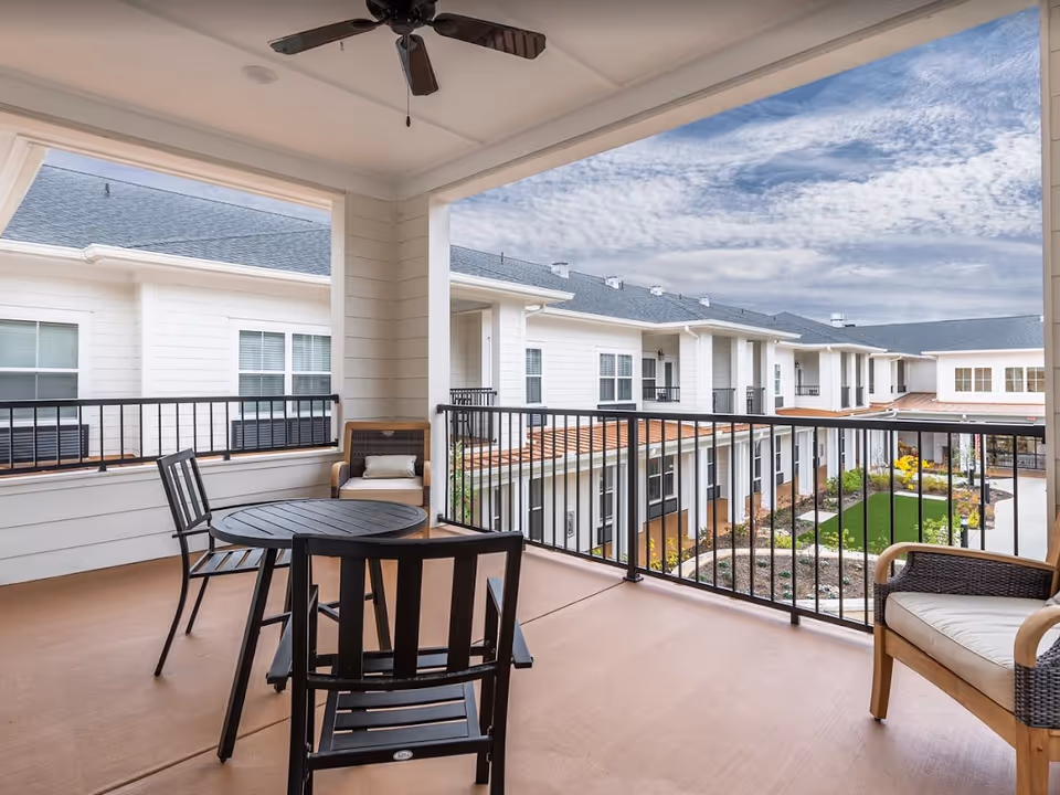 Covered outdoor balcony area with a round black table, three black chairs, and two cushioned armchairs. The balcony overlooks a courtyard with landscaped greenery and a multi-story building with white siding and multiple windows under a partly cloudy sky.
