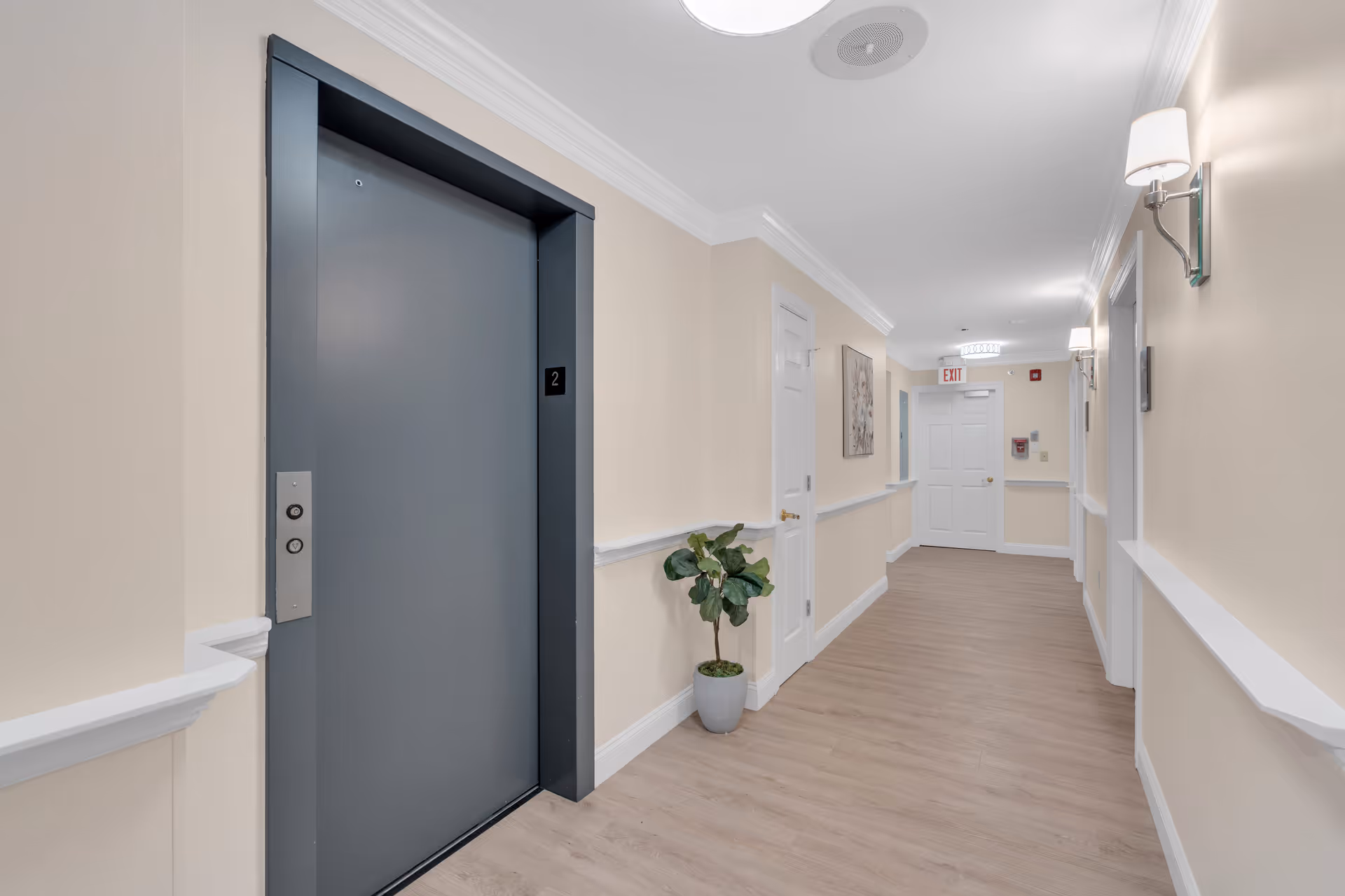 Interior hallway of a senior living facility with beige walls, light wood flooring, white trim, and a closed gray elevator door marked with the number 2. The hallway is decorated with a potted plant, wall sconces, and a framed picture, leading to a white exit door at the end.