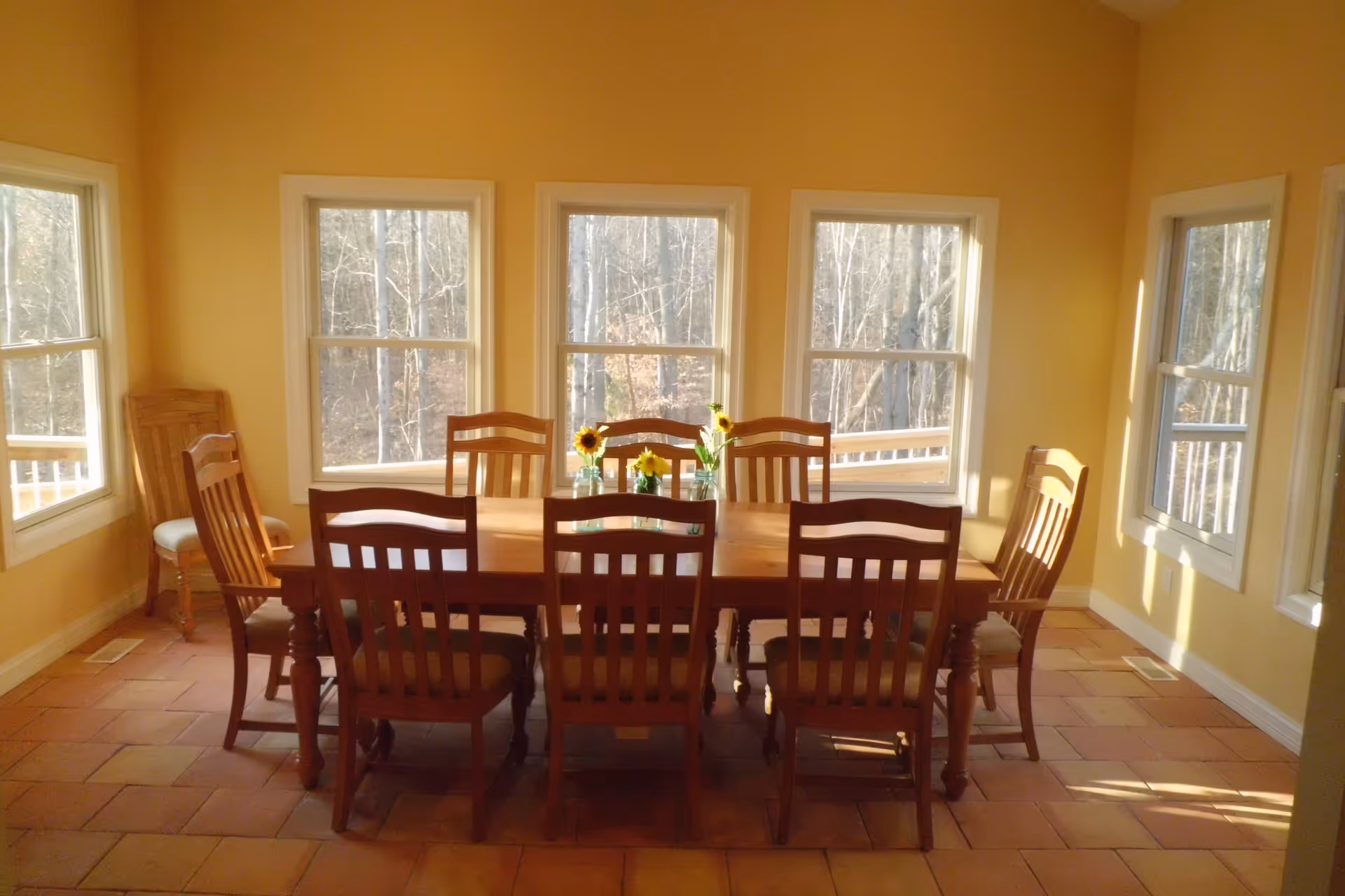 A bright dining room with a wooden dining table surrounded by eight wooden chairs. The room has multiple windows allowing natural light to fill the space, and small vases with sunflowers are placed on the table. The walls are painted a warm yellow color and the floor is tiled.