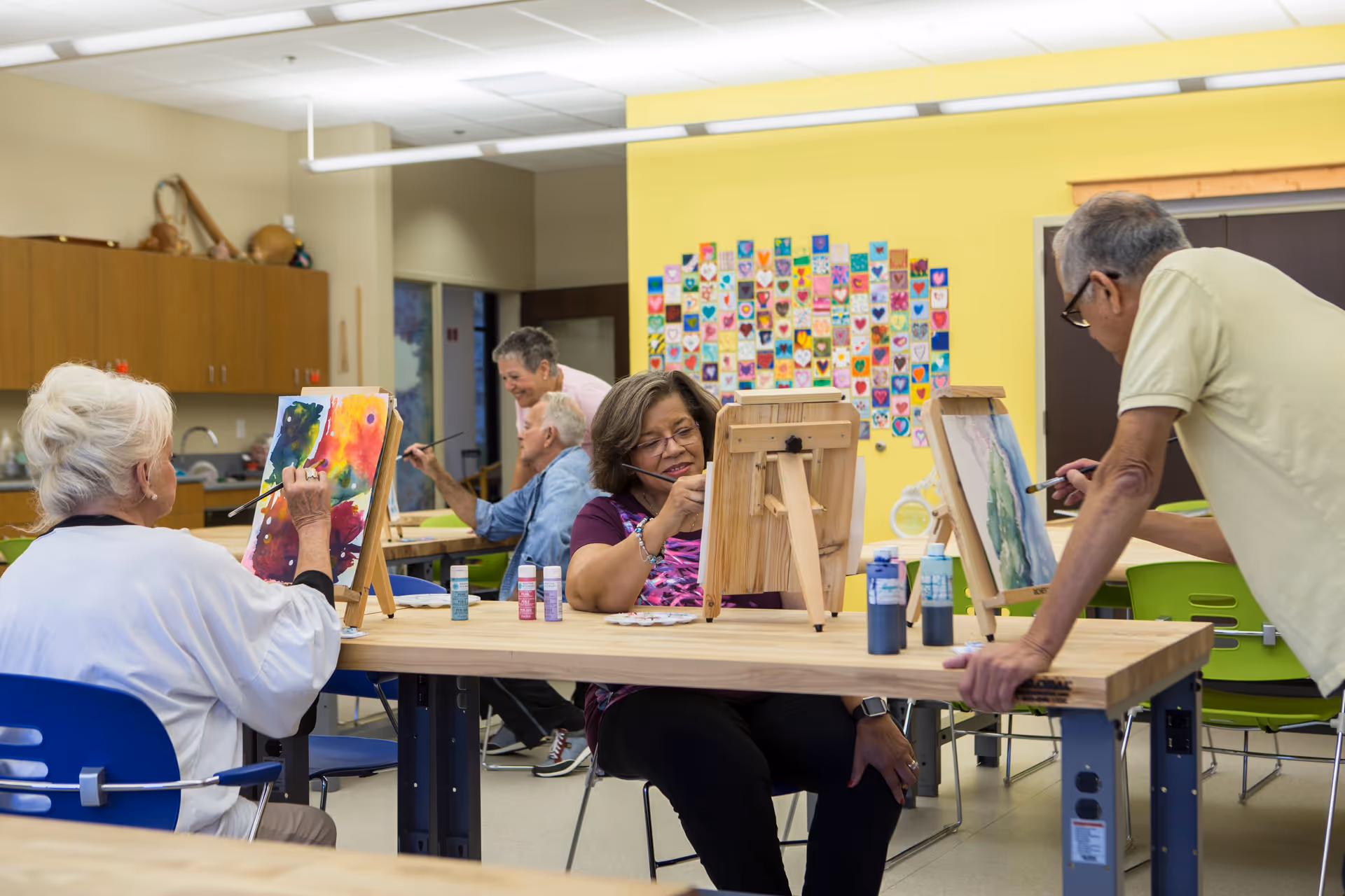 A group of elderly people engaged in a painting activity in a bright, spacious room. They are seated around tables with easels, paint bottles, and brushes, creating colorful artwork. The room has a yellow wall decorated with a collage of heart-shaped art pieces.