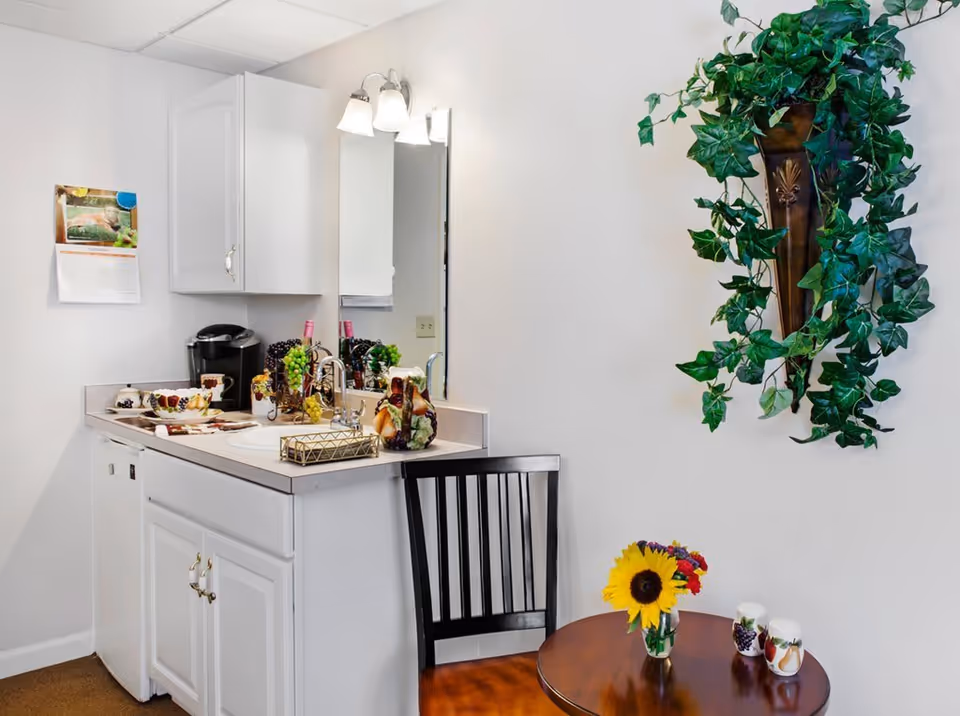 A small kitchenette area with white cabinets, a sink, and a coffee maker. There is a mirror above the sink with a light fixture. A black chair is placed next to a round wooden table with a small vase of sunflowers and other flowers, along with decorative salt and pepper shakers. A green leafy plant is mounted on the wall above the table.