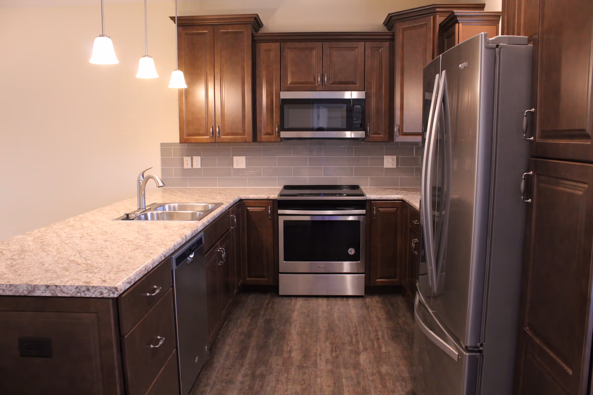 Modern kitchen with dark wood cabinets, stainless steel appliances including a refrigerator, oven, and microwave, a double sink, and a light-colored granite countertop. Three pendant lights hang above the counter.
